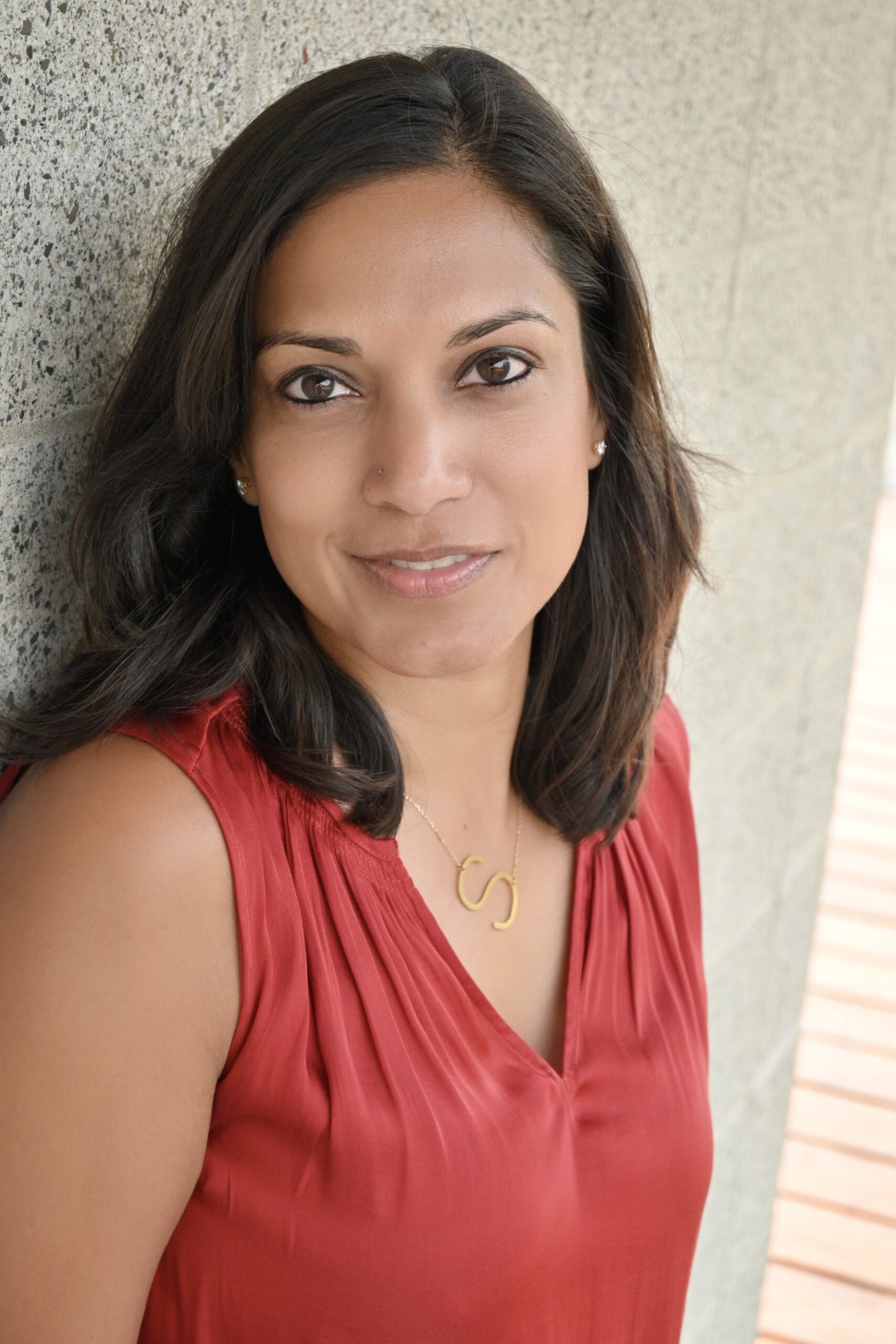 A headshot of Surita Jhangiani, a woman with shoulder-length dark brown hair, wearing an orange top. She is standing outdoors, looking at the camera with a semi-smile.