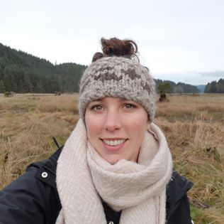 A headshot of Natasha Parrish, a woman standing on an estuary in her home Territory of Haida Gwaii. She is wearing a black jacket, scarf and wool headband. She is smiling in this photo.