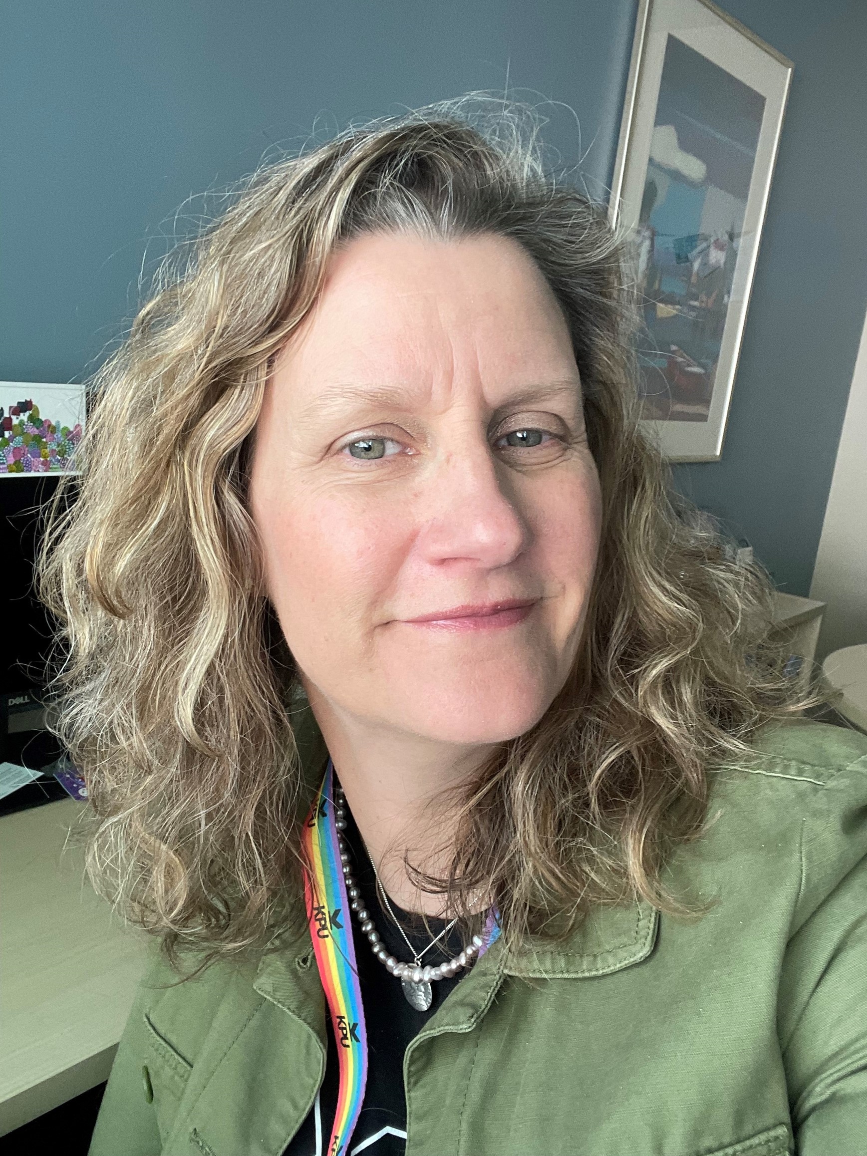 A headshot of Seanna Takacs, with brown wavy hair, wearing a green jacket, sitting in her KPU office and smiling at the camera.